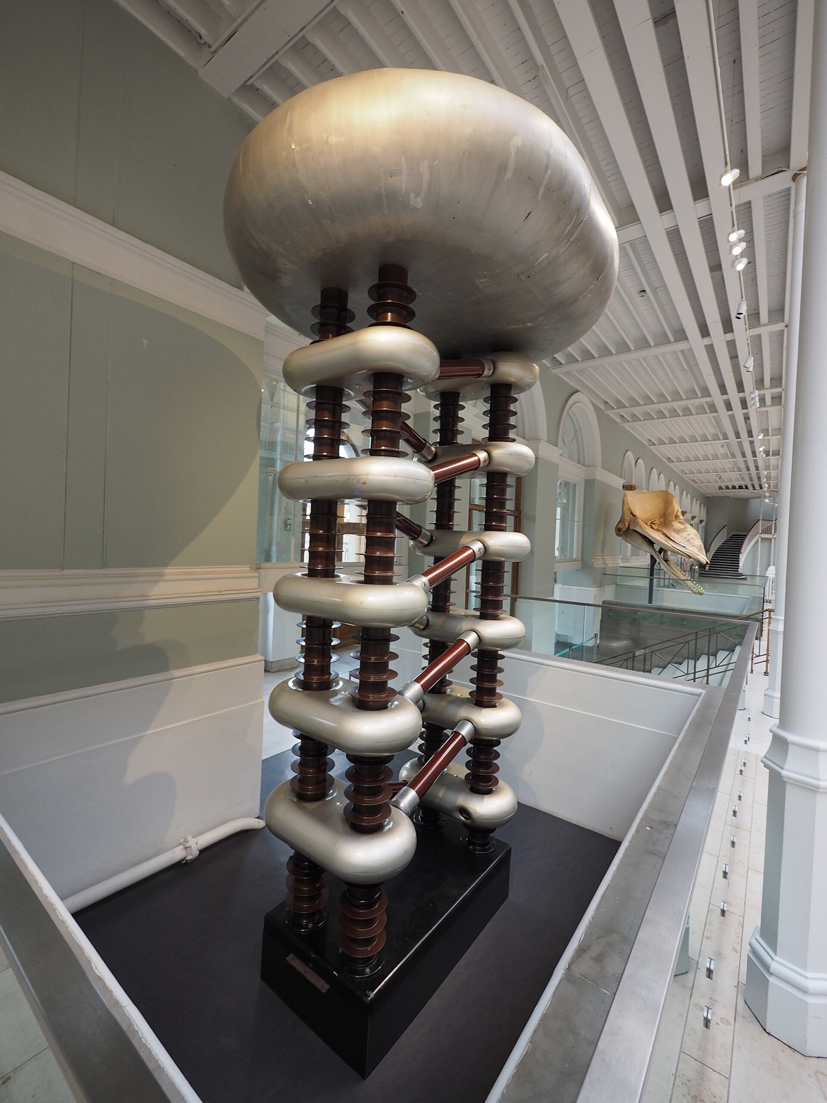 Tall Cockcroft–Walton voltage multiplier with repeated capacitor and rectifier stages inside a laboratory.
